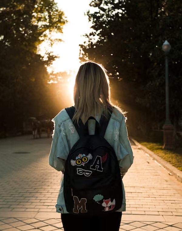Adolescente de mochila, de costas, caminhando num parque ao pôr do sol